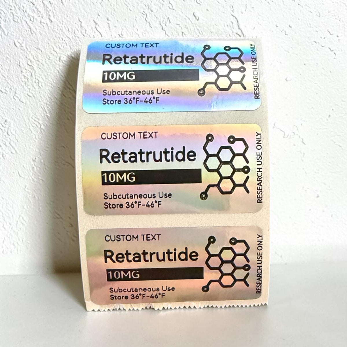 Three rectangular holographic labels featuring the text CUSTOM TEXT, Retatrutide, 10MG, Subcutaneous Use, Store 36°F-46°F, and RESEARCH USE ONLY. Each label includes a black graphic of a chemical molecular structure on the right side.