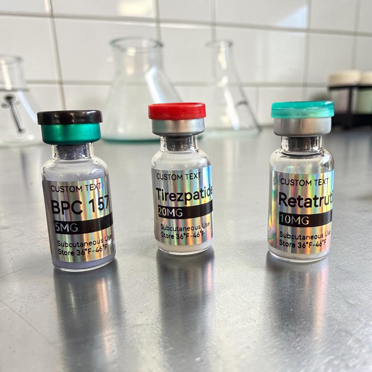 Three small glass vials with holographic labels. The left vial has a dark green cap and reads BPC 157 5MG. The middle vial has a red cap and reads Tirezpatide 20MG. The right vial has a teal cap and reads Retatrutide 10MG.