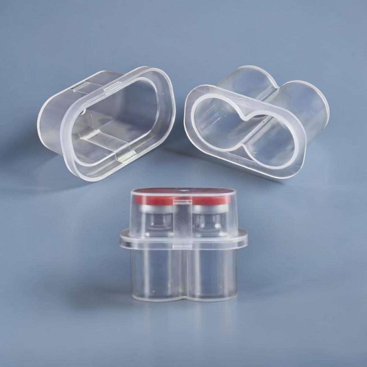 A set of clear plastic dual-vial holders with an oval-shaped rim. Two empty holders are positioned at the top, while a third holder at the bottom contains two small glass vials with red caps secured inside the transparent structure.