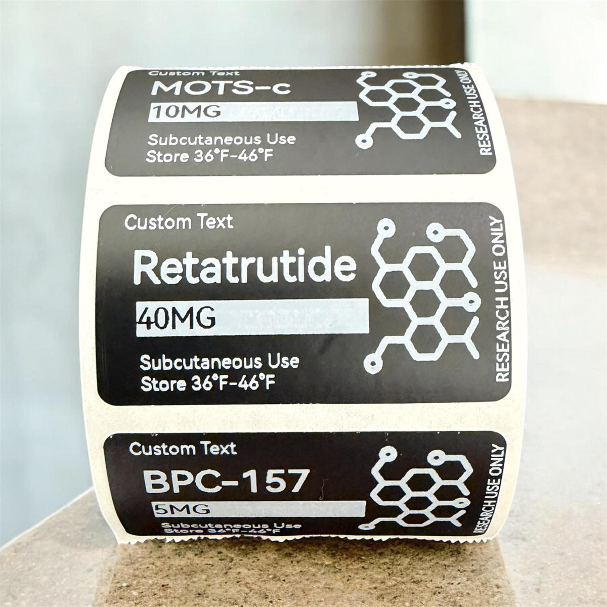 A roll of rectangular adhesive labels with a black background and white text. Each label features the text Custom Text, a chemical name like MOTS-c, Retatrutide, or BPC-157, dosage in MG, Subcutaneous Use, and Store 36°F-46°F.